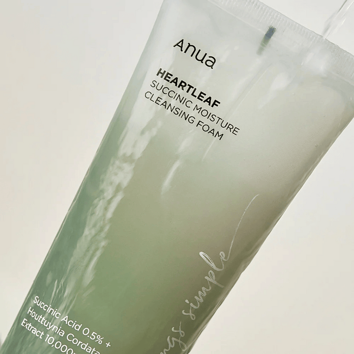 En genomskinlig flaska med Anua Heartleaf Succinic Moisture Cleansing Foam. Flaskan är fylld med ett grönt, molnigt skum och texten på framsidan listar aktiva ingredienser som succinsyra och Houttuynia Cordata-extrakt.