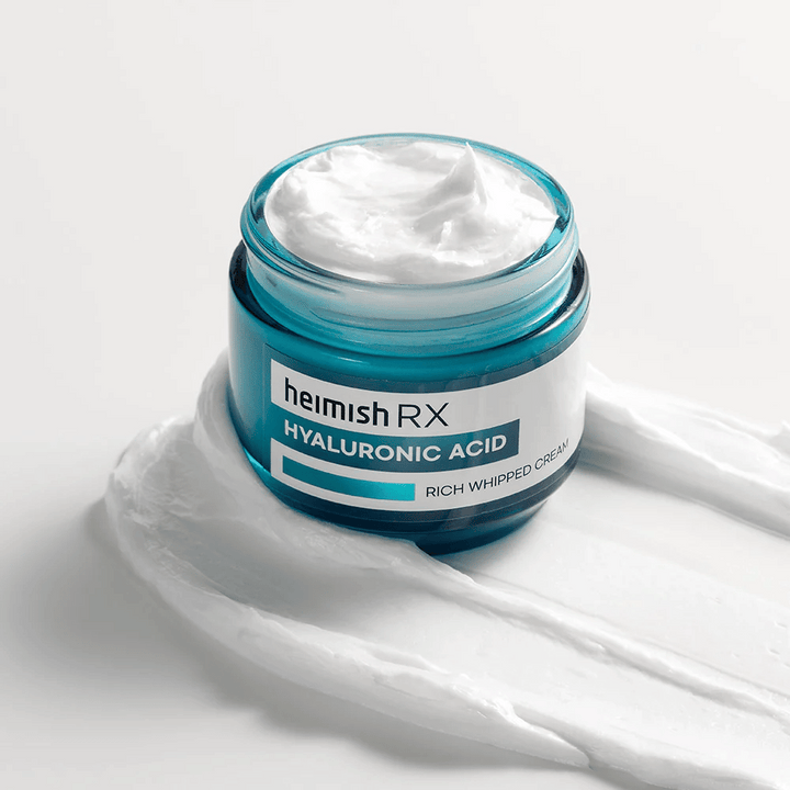 Heimish RX Hyaluronic Acid Rich Whipped Cream i en blå glasburk med rik och fluffig konsistens, utspridd på en vit bakgrund. Intensivt återfuktande ansiktskräm med hyaluronsyra för djup återfuktning och mjuk hud.