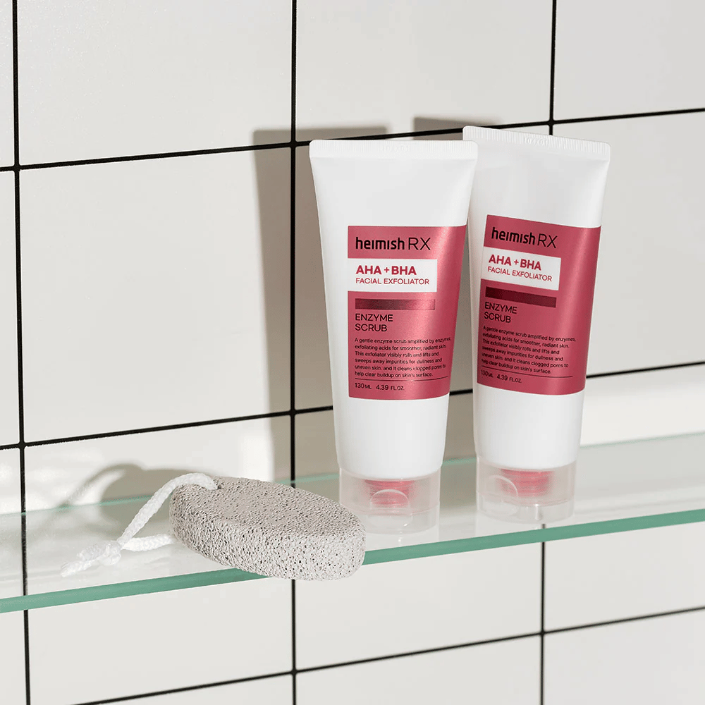 Två tuber Heimish RX AHA + BHA Facial Exfoliator placerade på en glashylla i ett badrum med vit kakelvägg. Bredvid produkterna ligger en pimpsten med ett vitt snöre. En stilren och minimalistisk hudvårdsbild.