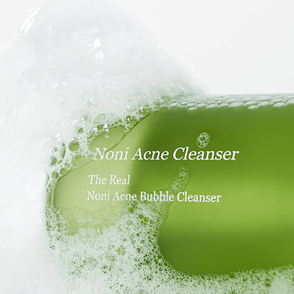 Flaskan med The Real Noni Acne Bubble Cleanser är i fokus och omges av rikligt med skum. Den friska gröna färgen signalerar naturliga ingredienser, perfekt för att rengöra och bekämpa akne. En idealisk produkt för en ren och klar hy.